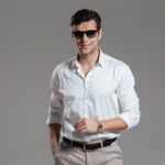 Portrait of a smiling young man dressed in white shirt wearing eyewear while standing and looking at camera isolated over gray background